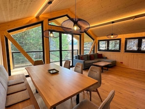 Ein geräumiges Zimmer mit Holztisch, Stühlen, einer Couch und großen Fenstern mit Blick ins Freie.
