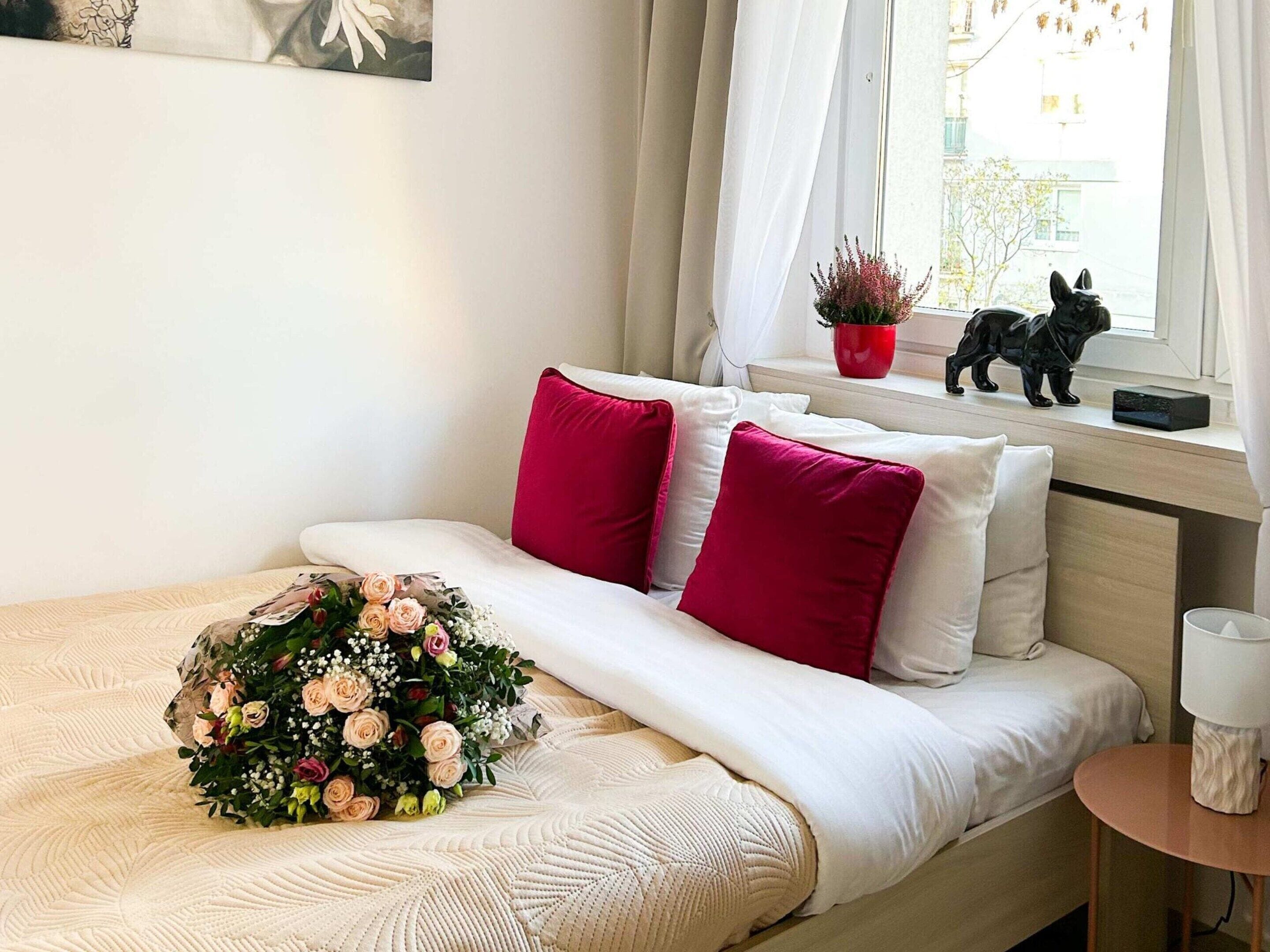 Een opgemaakt bed met bloemen, een raam met uitzicht op gebouwen, en een tafeltje met een lamp.