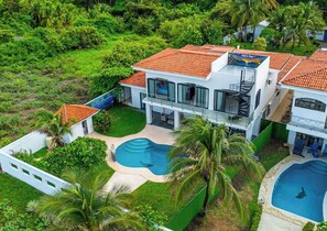 Ein modernes, zweistöckiges Haus mit Swimmingpool, umgeben von üppiger Vegetation und Palmen.