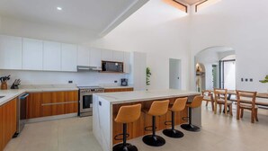 A modern kitchen with white cabinets, a central island with bar stools, and a dining area with wooden furniture. - Villa Bajamar - Costa Rica (Bajamar)