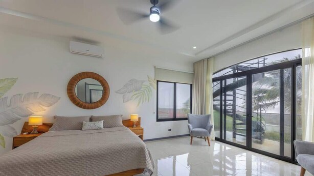 A bedroom with a bed, bedside lamps, a round mirror, a window with curtains, and a view of a staircase outside. - Villa Bajamar - Costa Rica (Bajamar)