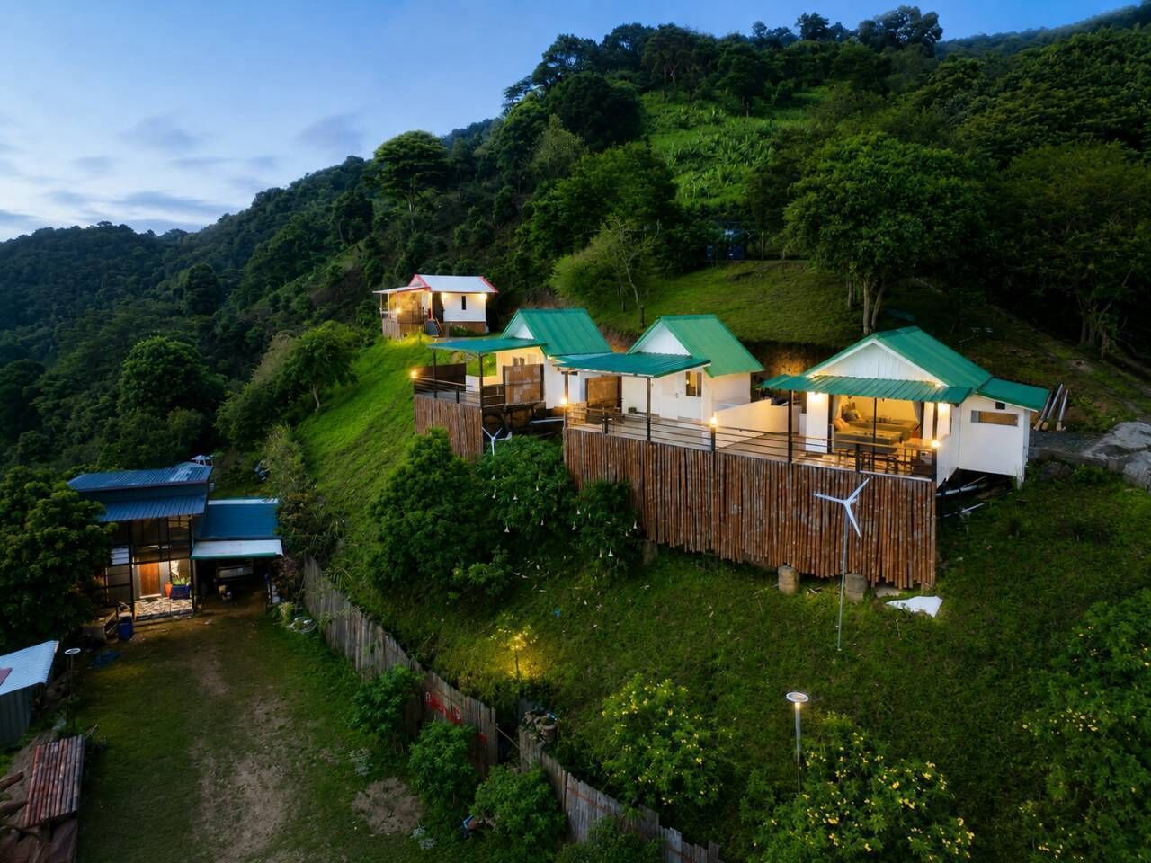 Una propiedad en una ladera con varias construcciones, incluyendo una casa principal con techo verde y una estructura más pequeña con techo blanco.