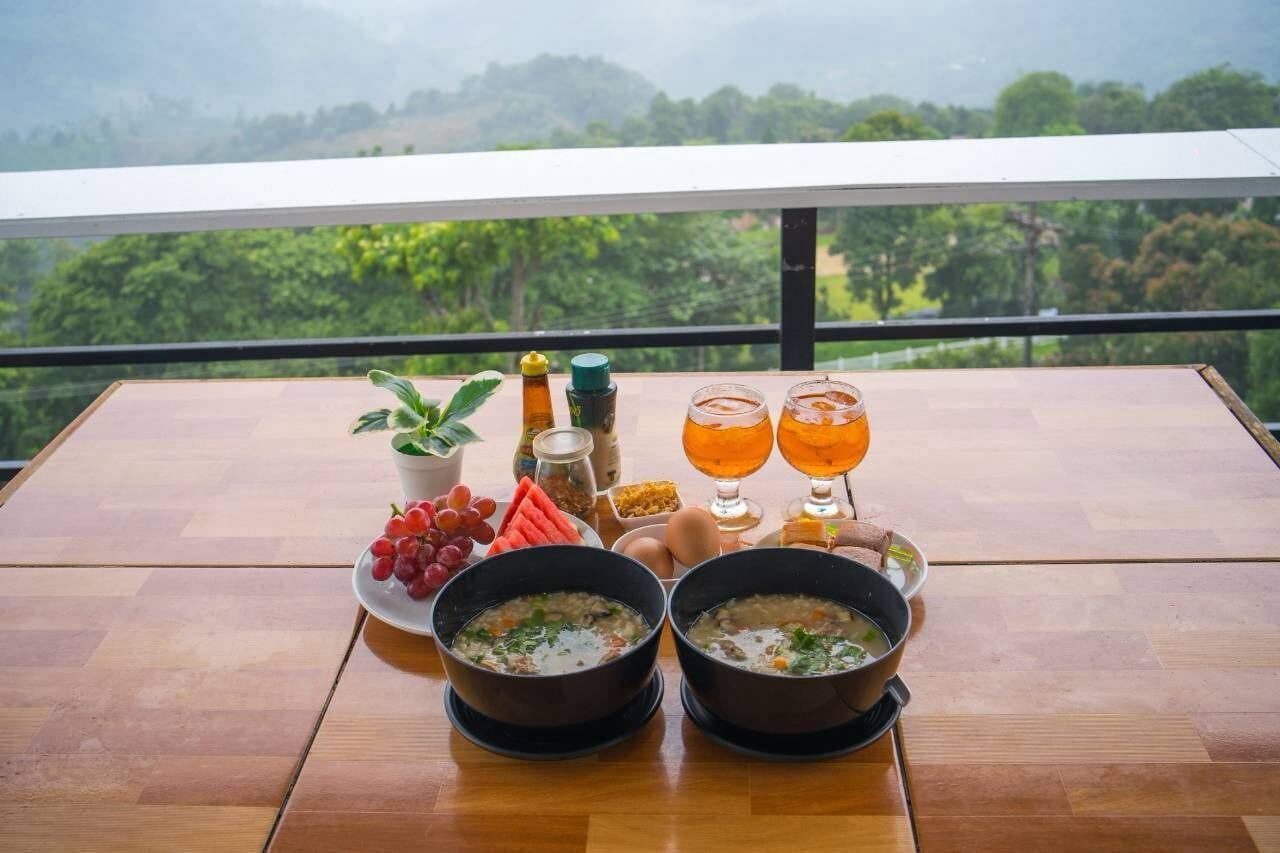 Una mesa de comedor con dos tazones de sopa, un plato de uvas, una botella de aceite y dos vasos de una bebida amarillenta, frente a una vista panorámica.