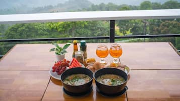 Una mesa de comedor con dos tazones de sopa, un plato de uvas, una botella de aceite y dos vasos de una bebida amarillenta, frente a una vista panorámica.