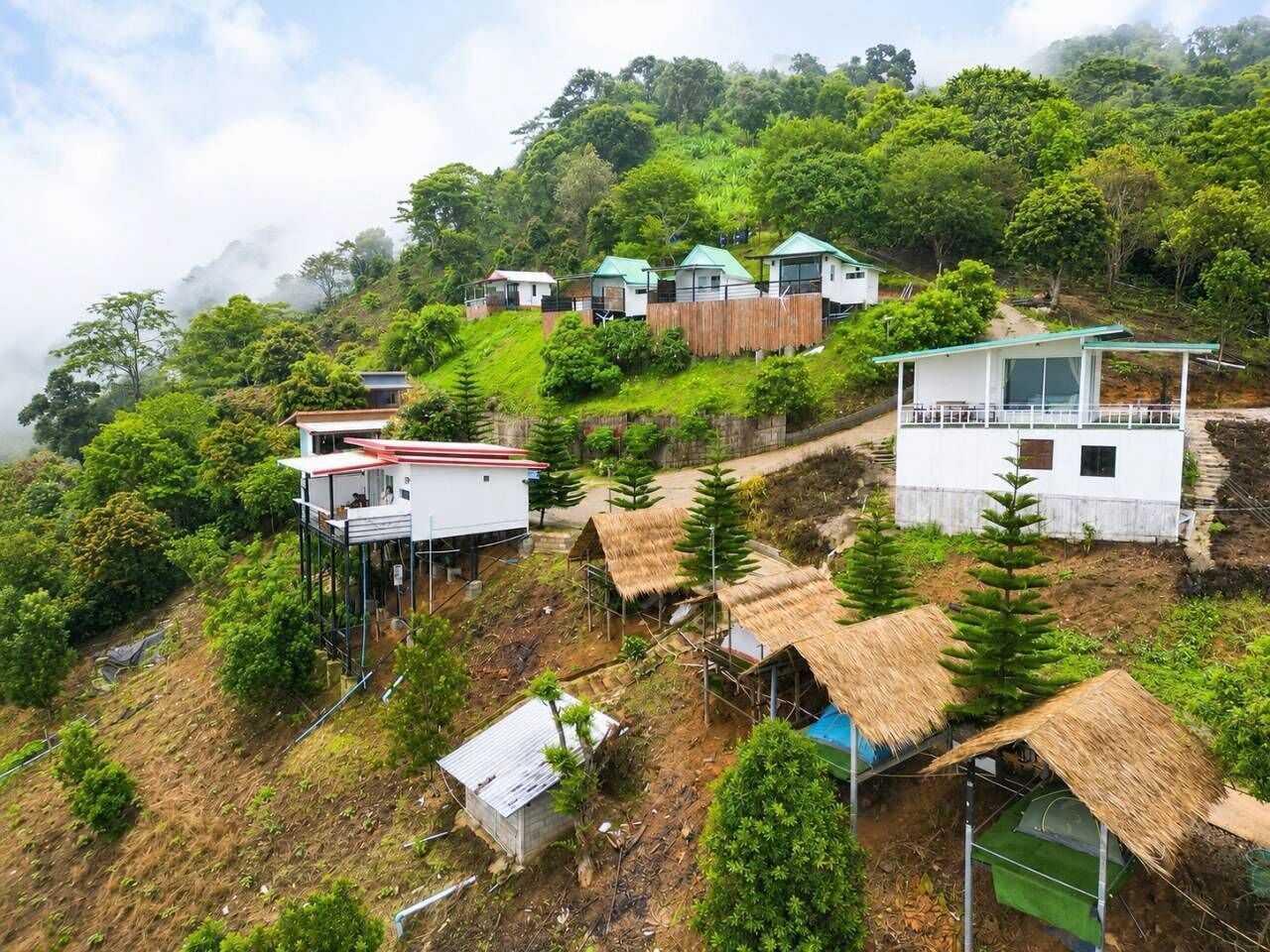 A hillside settlement with multiple buildings, some with thatched roofs and others with modern architecture, surrounded by lush greenery.