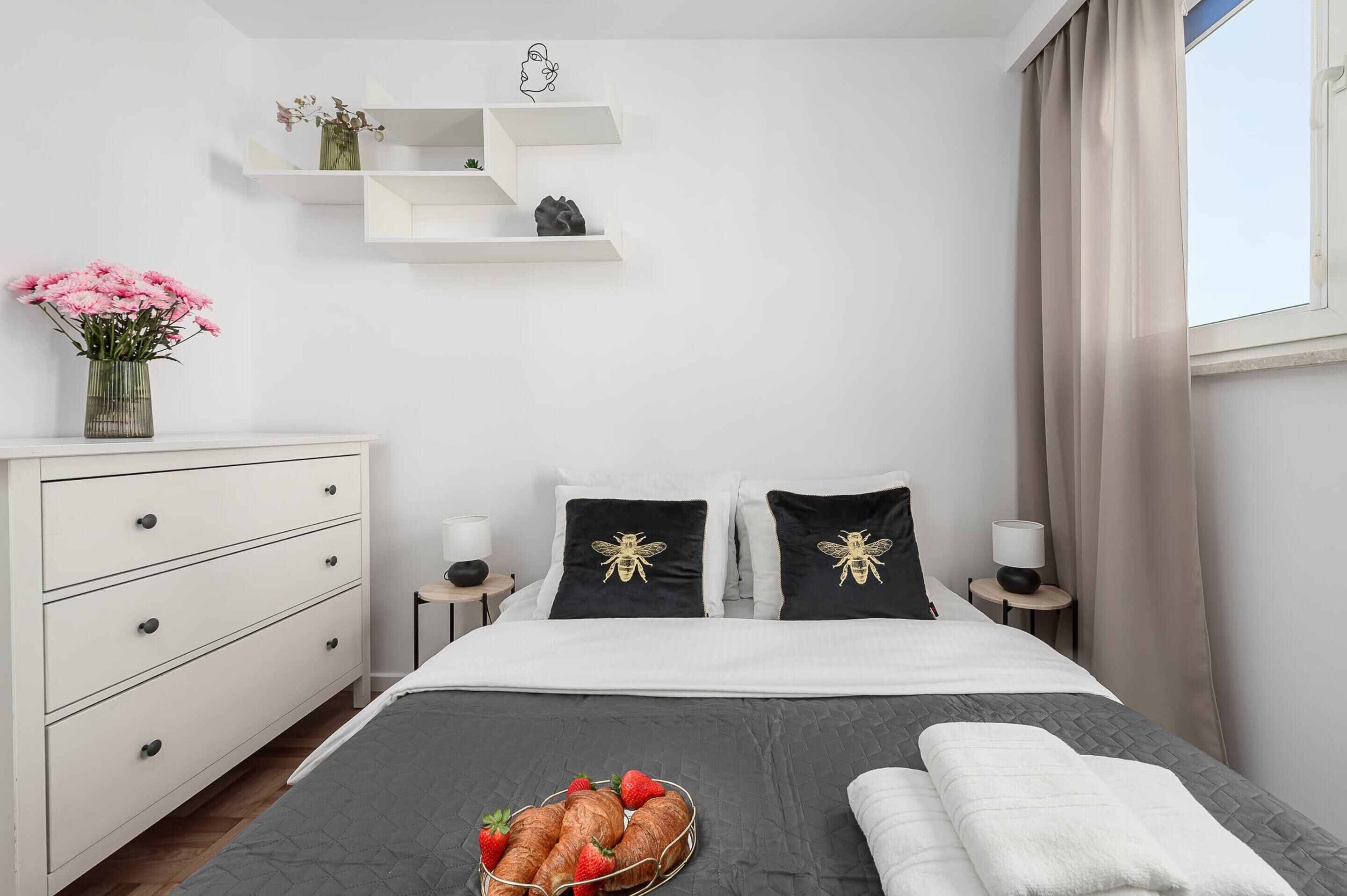 Een hotelkamer met een bed, nachtkastjes, een dressoir, een vaas met bloemen en een raam met gordijnen.