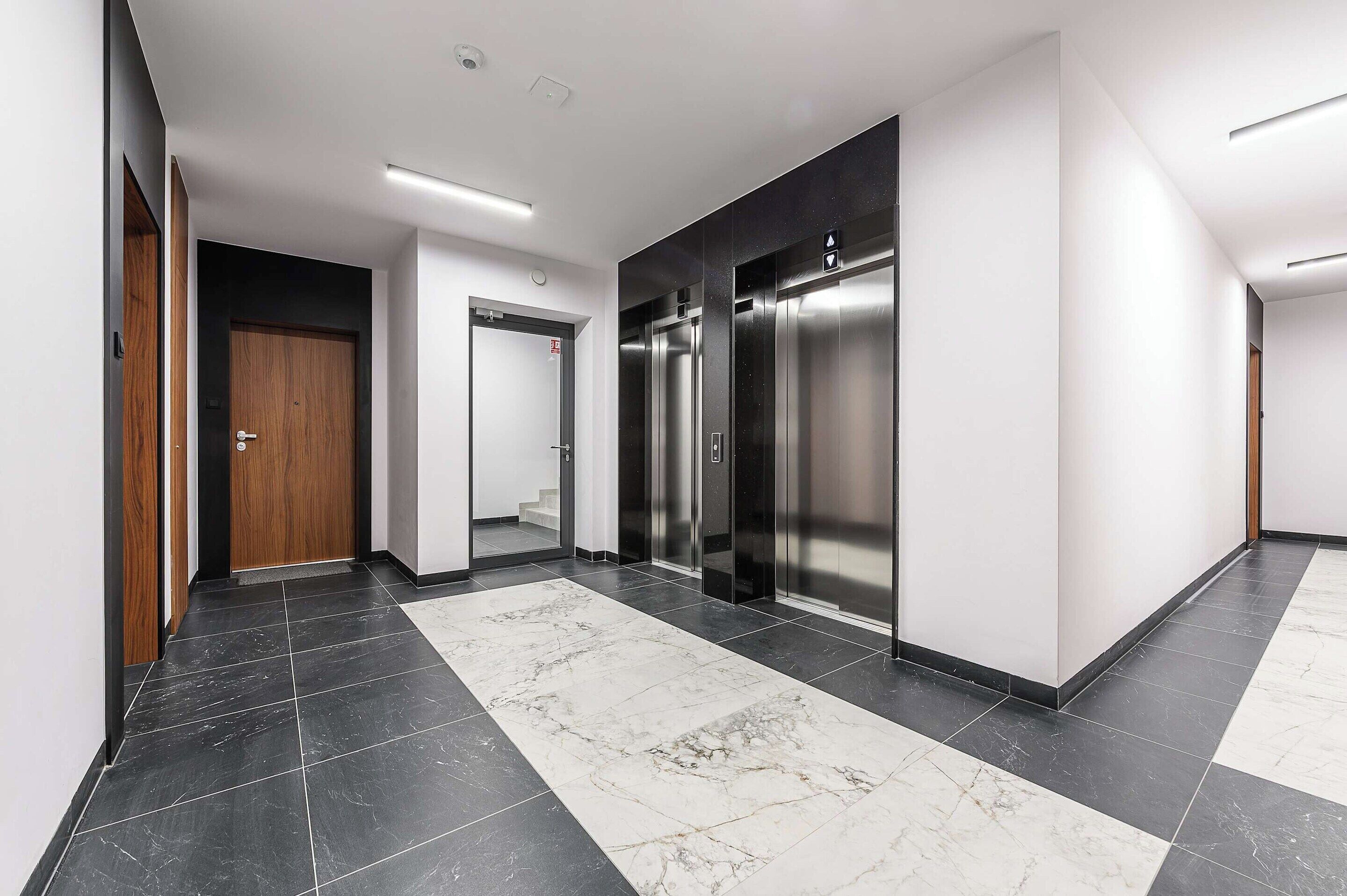 A modern hallway with a marble floor, wooden doors, and two elevators.