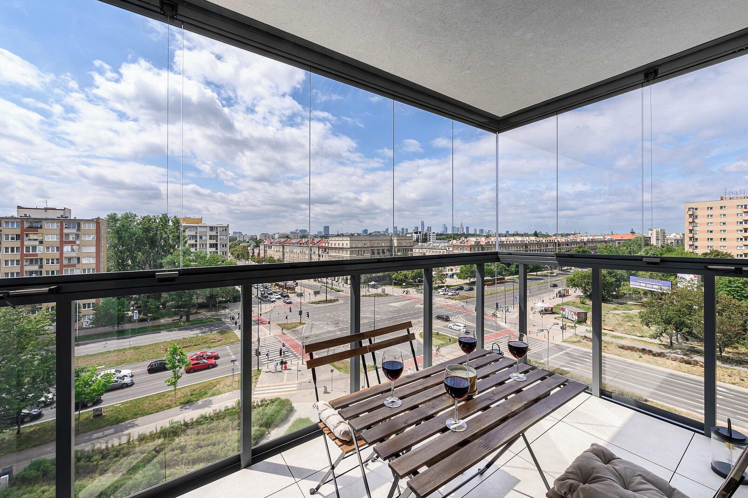 A balcony with a view of a cityscape, a wooden bench, and two glasses of red wine.