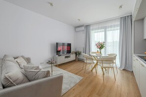 A modern living room with a sofa, a TV displaying 'NETFLIX', a dining area with a table and chairs, and a kitchen with cabinets. - Family Home Steps From the Beach (Warsaw)