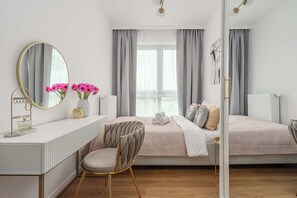 A bedroom with a white dresser, a vanity with a mirror, a chair, two beds with bedding, and a window with curtains. - Family Home Steps From the Beach (Warsaw)