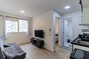 A modern living room with a sofa, a TV on a stand, a kitchen area with a sink, and a view of a building through the window. - Studio in downtown São Paulo. Metro República. Easy access to Brás and 25 de Março. (São Paulo)