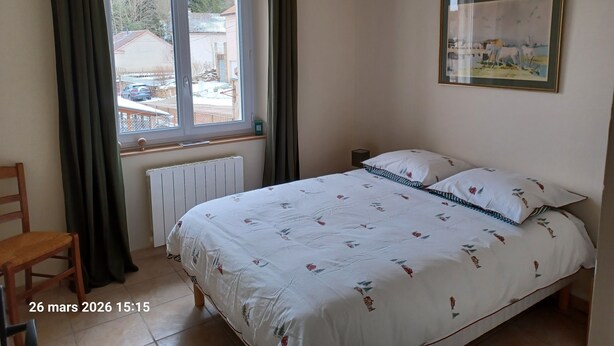Un dormitorio con cama, una silla, un ventanal con cortinas, un radiador y un cuadro en la pared.