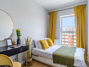 Ein Schlafzimmer mit Bett, Schreibtisch, Stuhl, einem Fenster mit Vorhängen und Blick auf Gebäude.