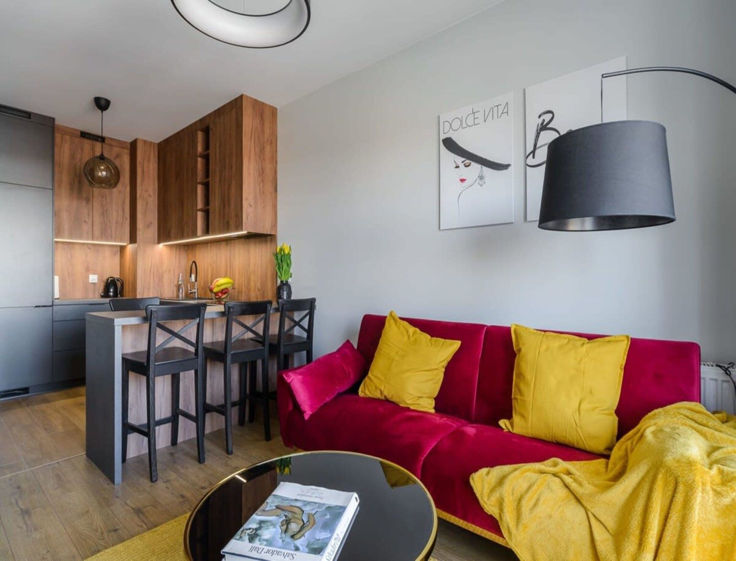 Ett modernt vardagsrum med en röd soffa, svarta matstolar, ett köksområde med träskåp och en hängande lampa.