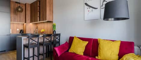 Un espacio de estar moderno con un sofá rojo, sillas de comedor negras, una zona de cocina con armarios de madera y una lámpara colgante.