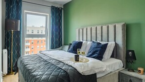 A bedroom with a bed, a nightstand, a lamp, and a view of a building through a window. - Quiet City Apartment for 4 (Łódź)