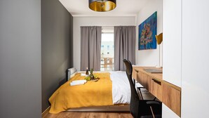 A modern hotel room with a bed, a desk, a chair, a painting, and a view of buildings outside. - City View Apartment in Warsaw (Łódź)