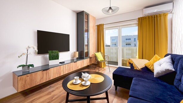 A modern living room with a flat-screen TV, a round coffee table, a sofa, and a balcony with yellow curtains. - City View Apartment in Warsaw (Łódź)