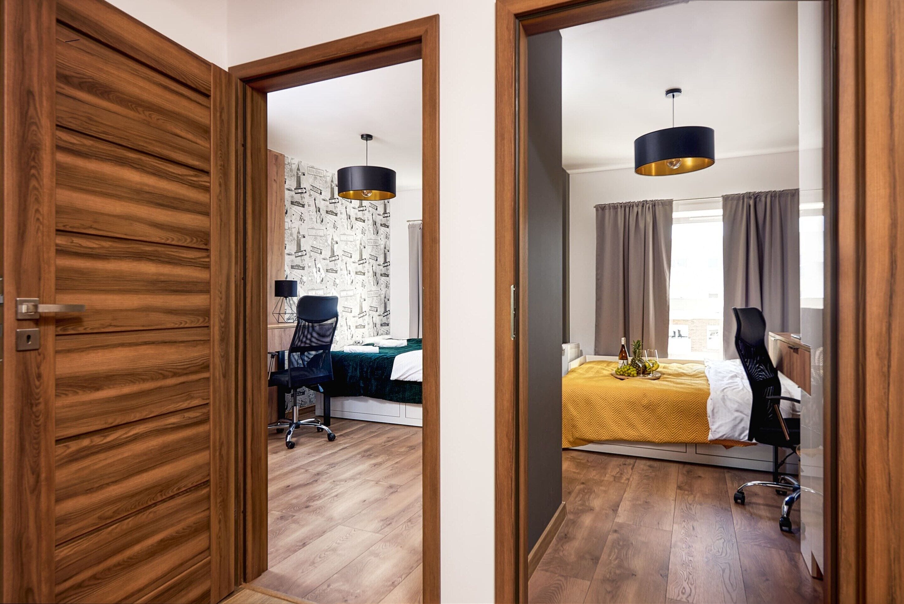 Une chambre d’hôtel avec un plancher en bois, un lit recouvert d’une courtepointe jaune, un bureau avec une chaise, un mur orné d’un papier peint représentant un paysage urbain et un luminaire suspendu moderne.