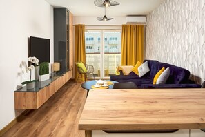A modern living room with a wooden dining table, a purple sofa with yellow cushions, and a view of the city through large windows. - City View Apartment in Warsaw (Łódź)