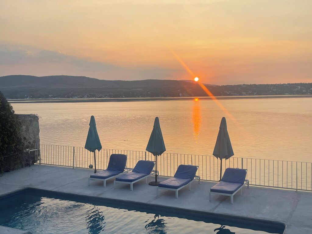 Un espace aménagé au bord de la piscine, avec des chaises longues et des parasols, offrant une vue sur une étendue d’eau au coucher du soleil.