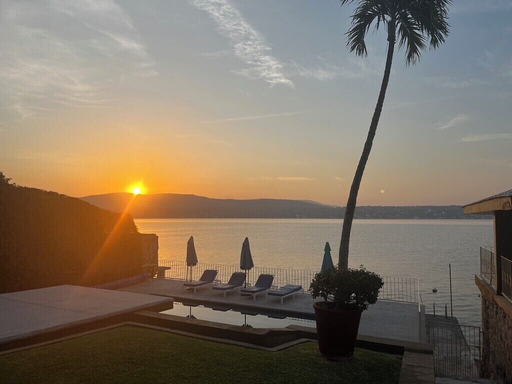 A poolside view with lounge chairs and umbrellas, overlooking a body of water during sunset.