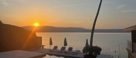 Vista de la piscina con sillones de descanso y sombrillas, con vista a un cuerpo de agua al atardecer.
