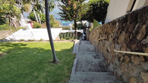 Unos escalones de concreto conducen a un muro de piedra, con una zona verde y árboles al fondo.