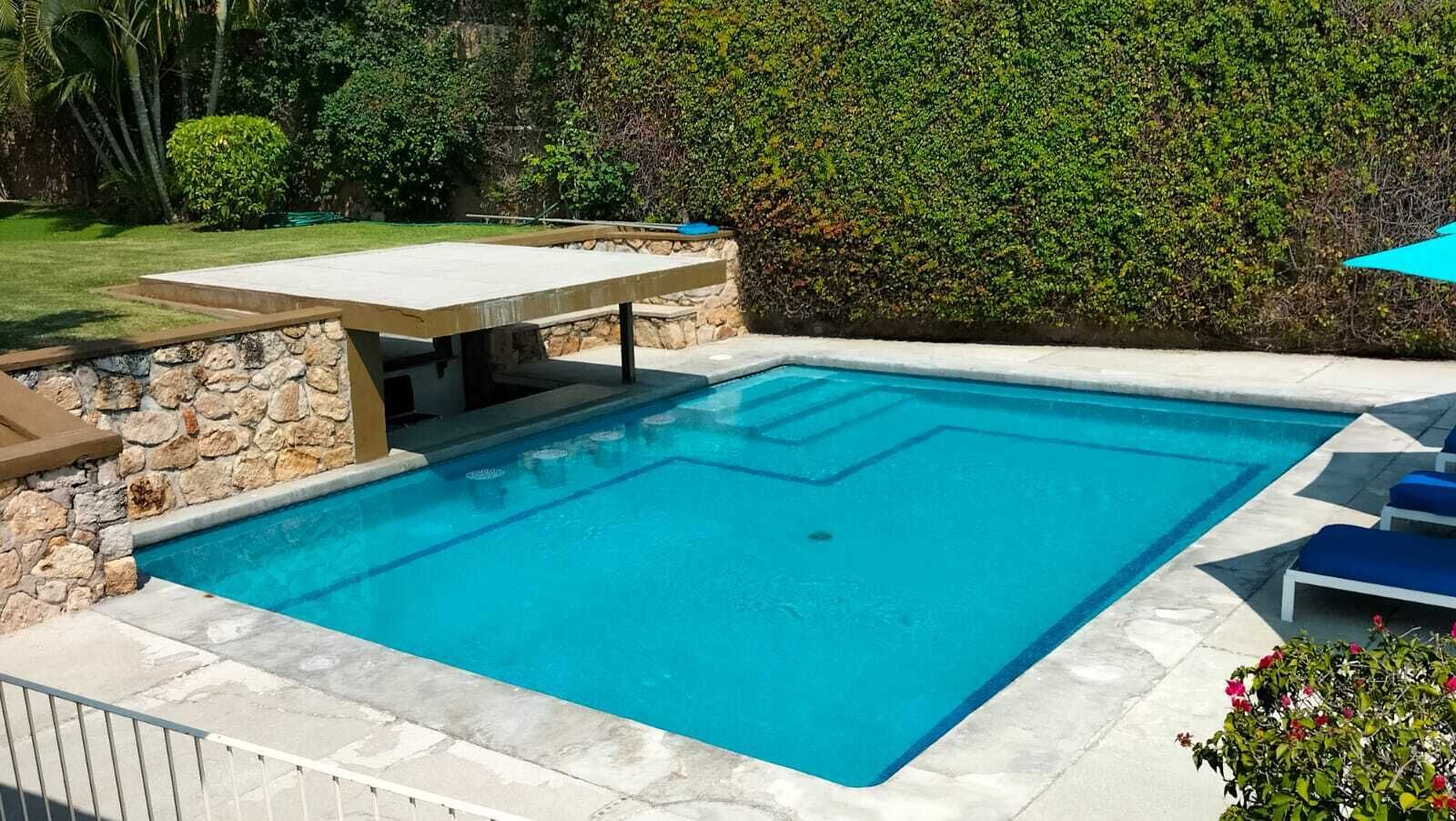 A rectangular swimming pool with a stone border, surrounded by a stone wall and a hedge.