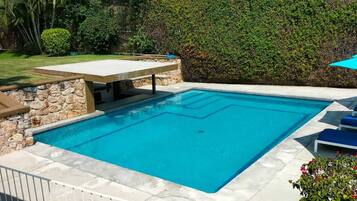 A rectangular swimming pool with a stone border, surrounded by a stone wall and a hedge.