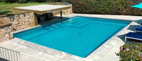 A rectangular swimming pool with a stone border, surrounded by a stone wall and a hedge.