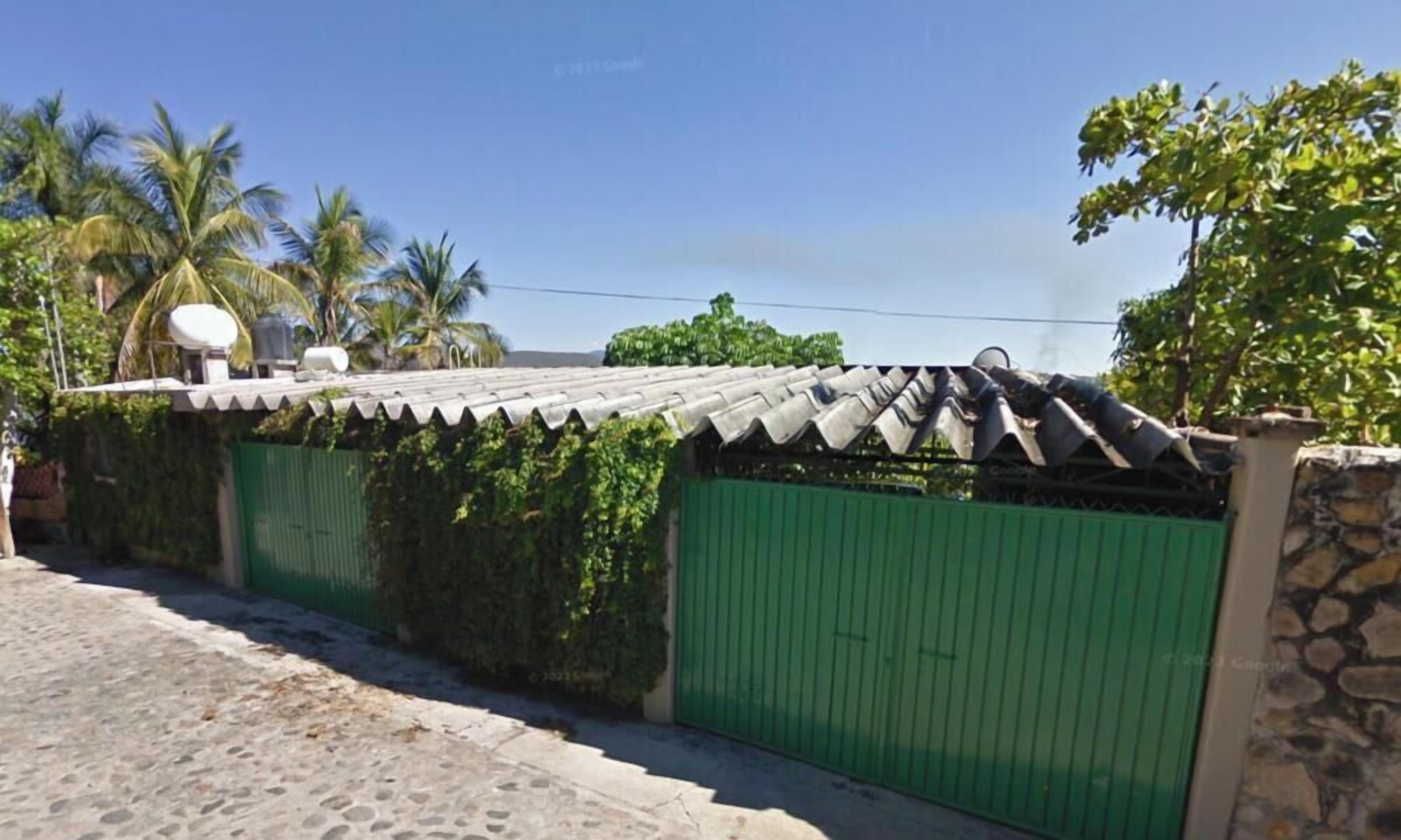 A gated property with a tiled roof and a green gate, surrounded by greenery and palm trees.