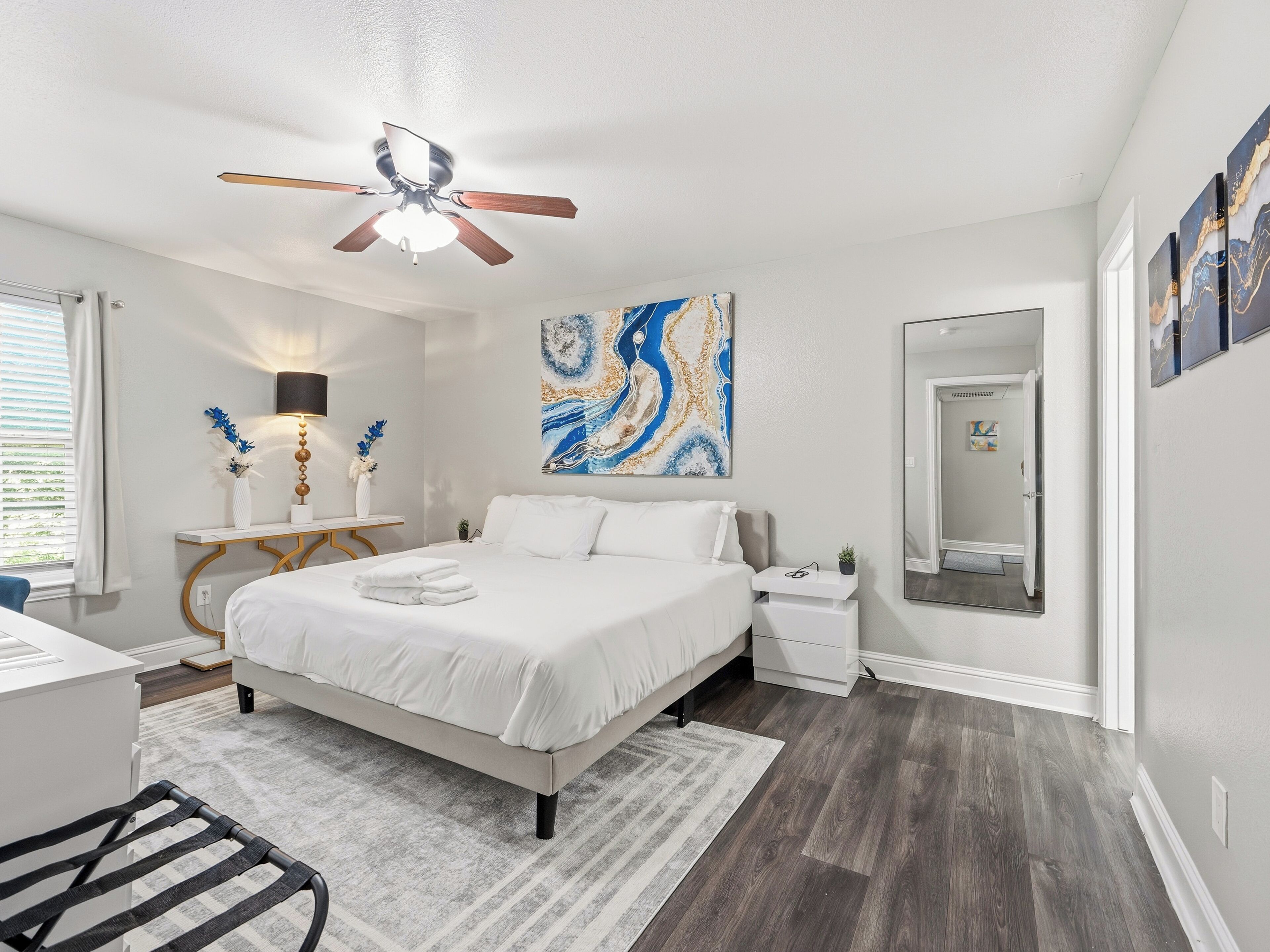 A bedroom with a bed, bedside tables, a ceiling fan, a large abstract painting, and a mirror.