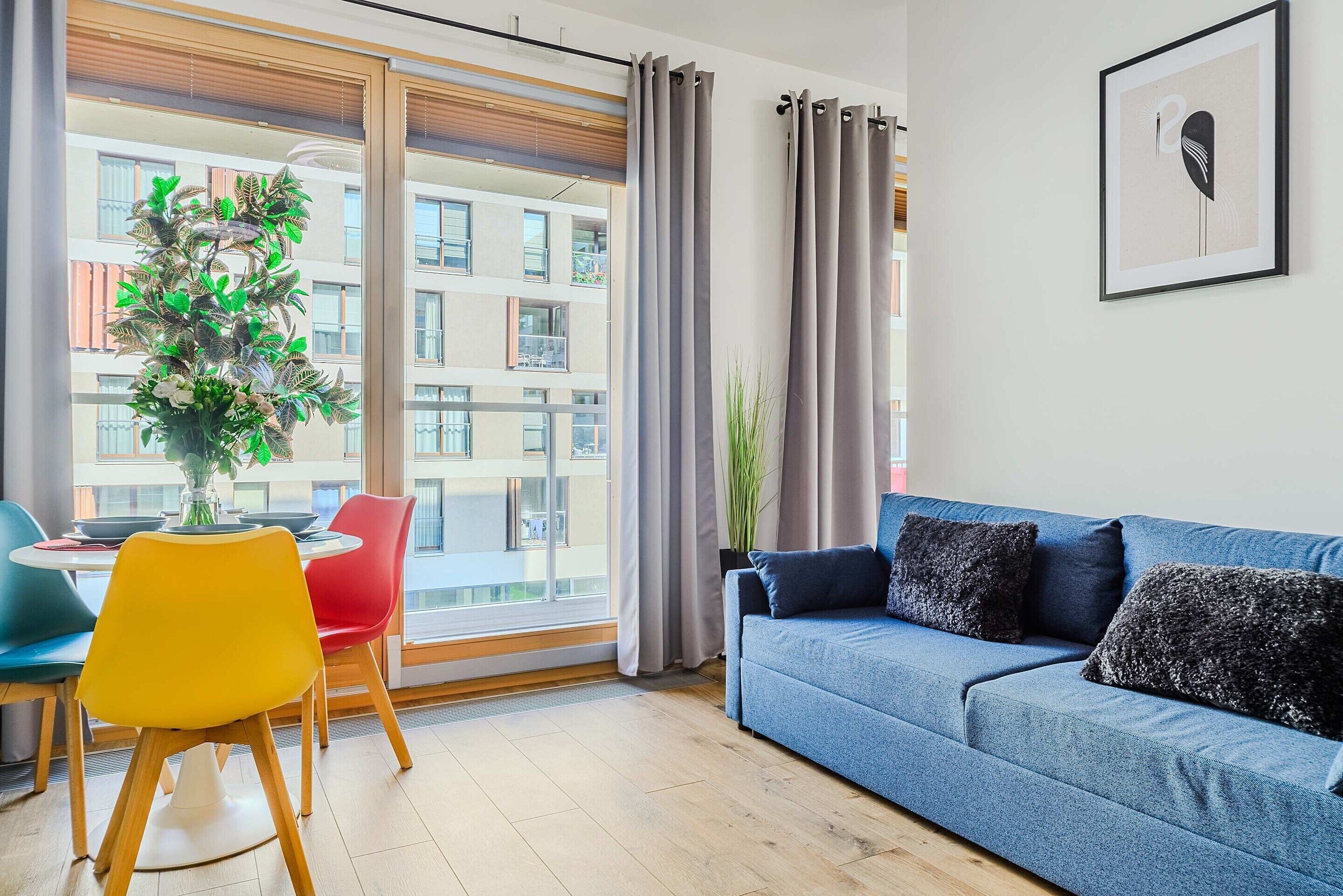 A modern living room with a blue sofa, a dining table with chairs, and a view of a building through large windows.