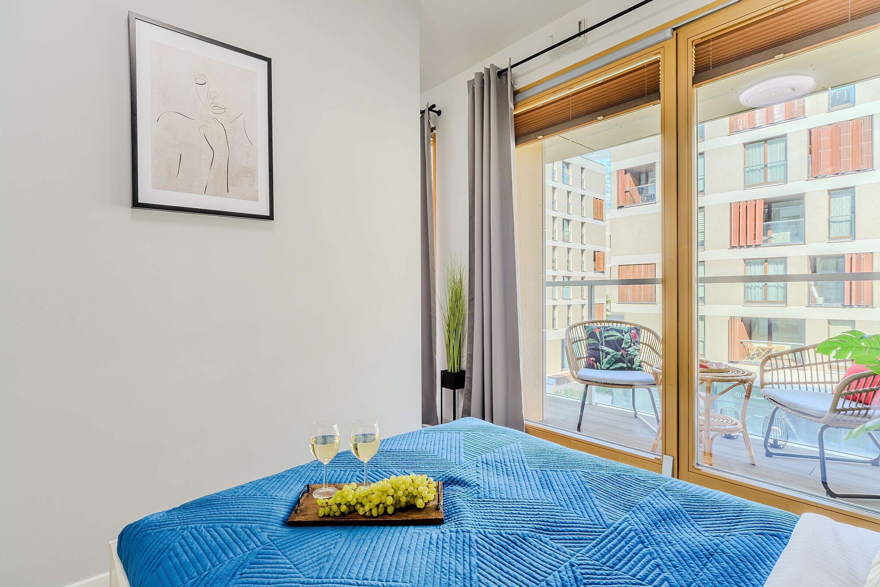 Quarto de hotel com uma cama, uma obra de arte emoldurada na parede, uma varanda com vista para edifícios e uma mesa com uma bandeja de uvas e copos.