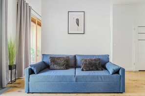 A blue sofa with two pillows, a framed abstract artwork on the wall, and a potted plant beside the window. - Stay Near Railway Hub (Warsaw)