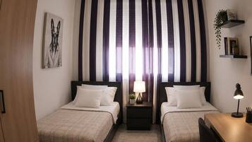 A hotel room with two beds, a desk, a nightstand, a lamp, and a large window with striped curtains.