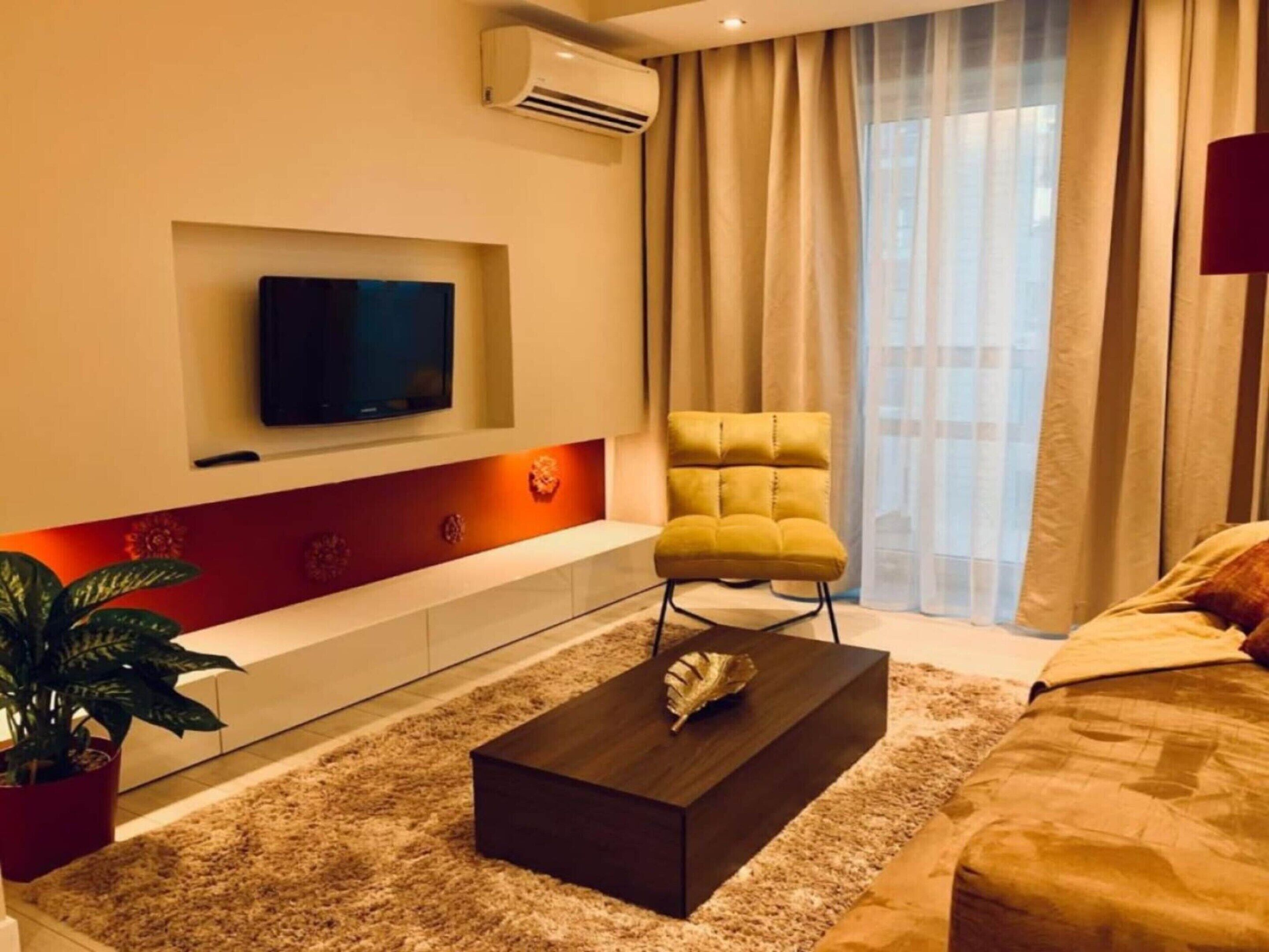 A modern living room with a flat-screen TV mounted on the wall, a yellow armchair, a dark coffee table, and a large window with curtains.