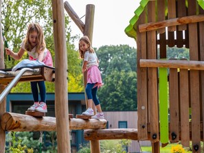 Two children playing on a wooden play structure with a slide and climbing elements.