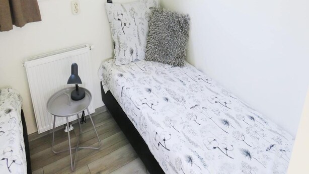 Quarto pequeno com uma cama de solteiro, radiador, escrivaninha com luminária e janela com cortinas.