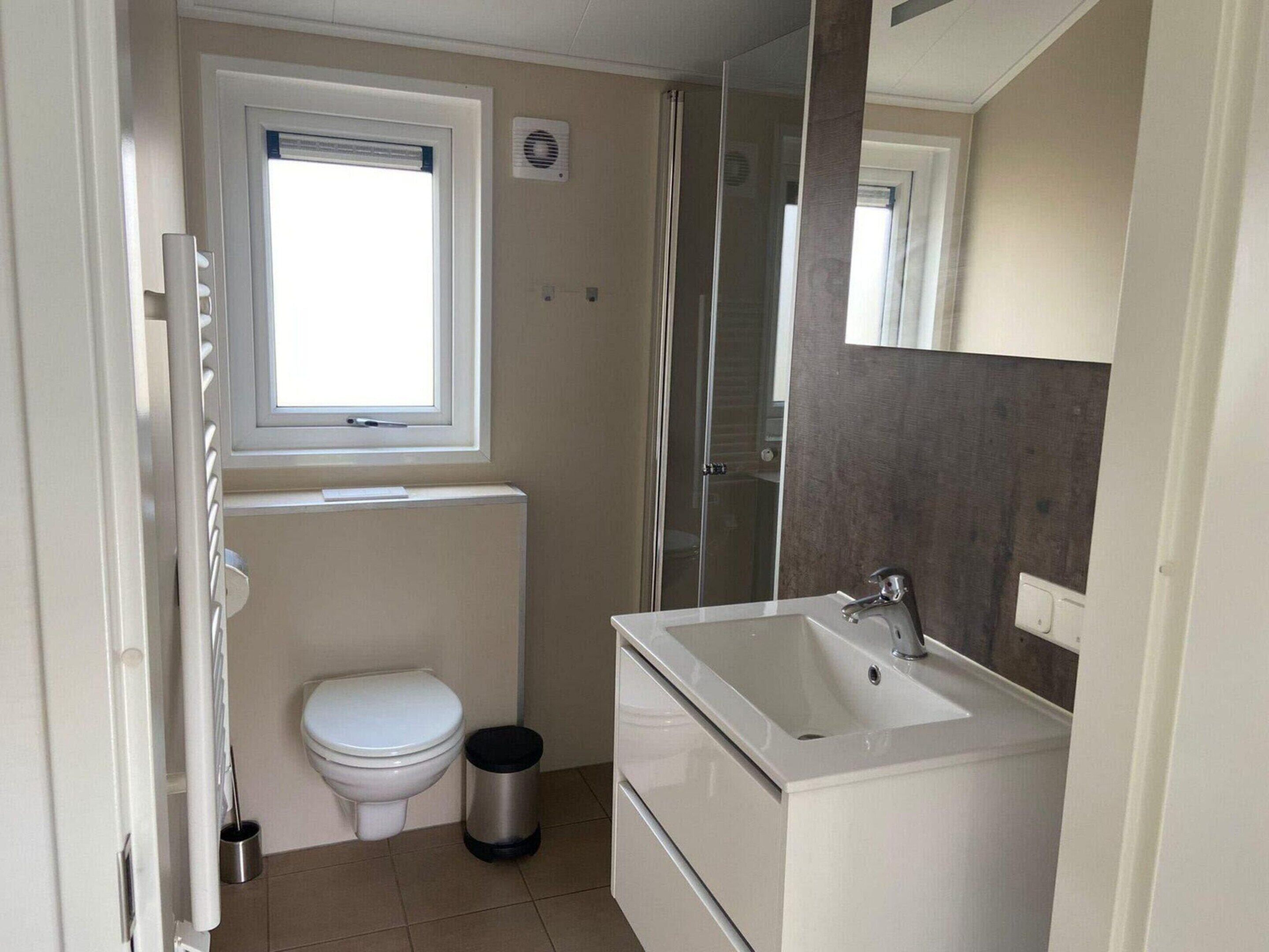 A compact bathroom with a toilet, a small trash bin, a window, and a shower area.