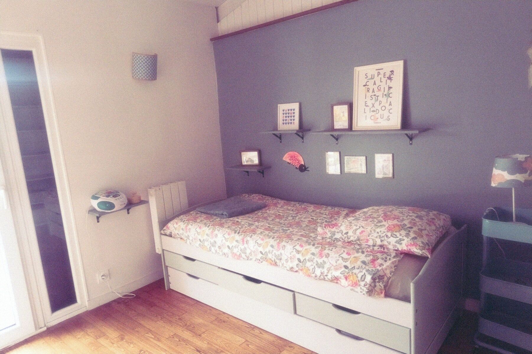 Une chambre à coucher avec un lit, une petite étagère avec des photos, une table de chevet avec une lampe et une fenêtre.