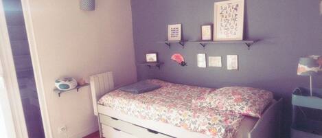 A bedroom with a bed, a small shelf with pictures, a bedside table with a lamp, and a window.