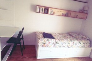 Ein kleines Zimmer mit einem Einzelbett, einem Schreibtisch und einem Stuhl. Über dem Bett befindet sich ein Regal mit Büchern und Dekoration.
