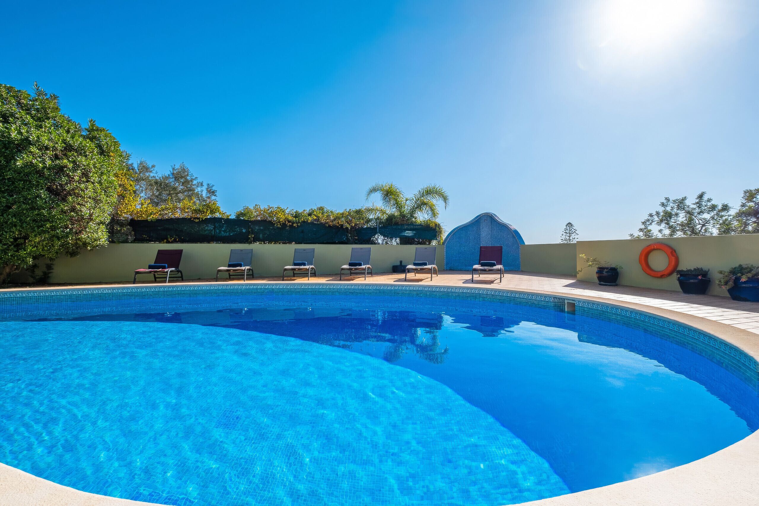 A clear blue swimming pool with lounge chairs and a sunshade, surrounded by greenery and a clear sky.