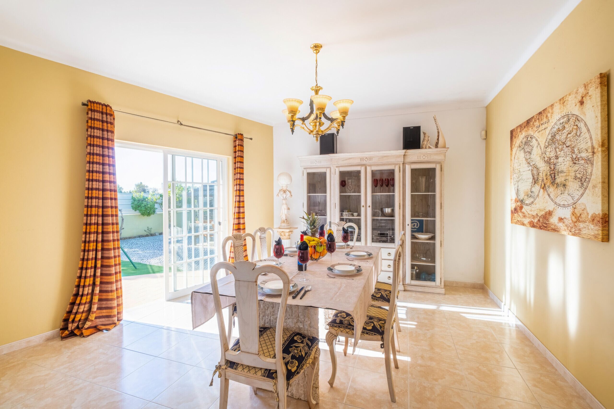 A dining room with a table set for four, chairs, a chandelier, a cabinet with glass doors, and a large world map on the wall.