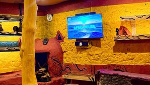 Een kamer met een aan de gele muur bevestigde televisie, een houten sculptuur en een stenen bank.