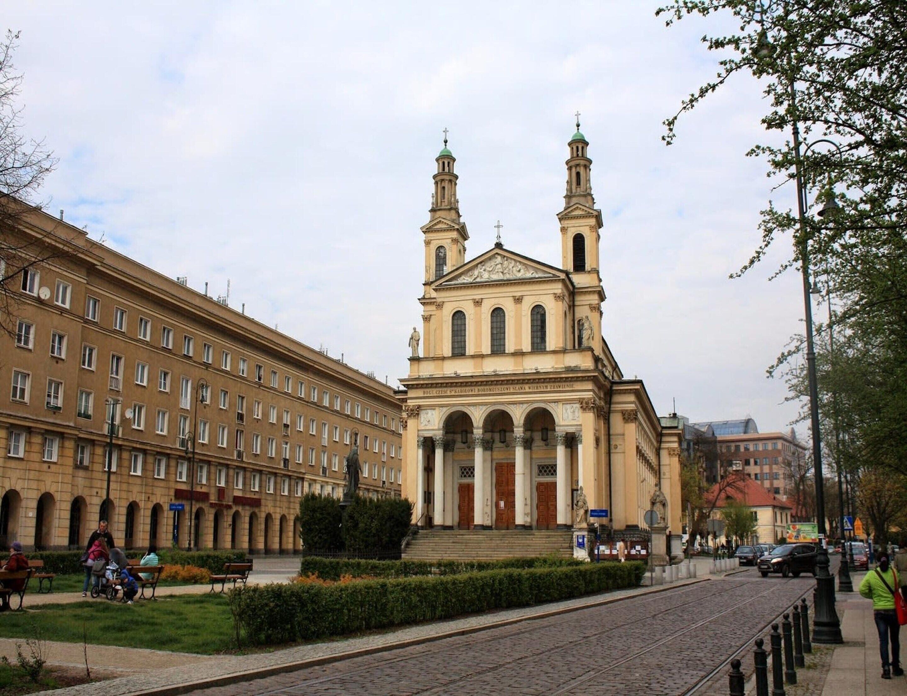 A historic building with a central dome and two towers, surrounded by a paved square with benches and a statue.