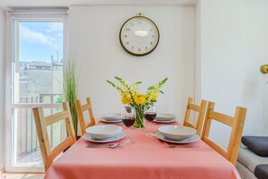 A dining area with a table set for two, a clock on the wall, and a view of buildings through a window. - Apartment With Private Balcony (Warsaw)