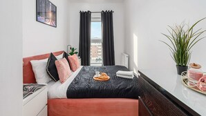 A hotel room with a bed, a bedside table with a plant, a window with curtains, and a view of buildings outside. - Beautiful Seaview Flat in Stagadon Island (Łódź)